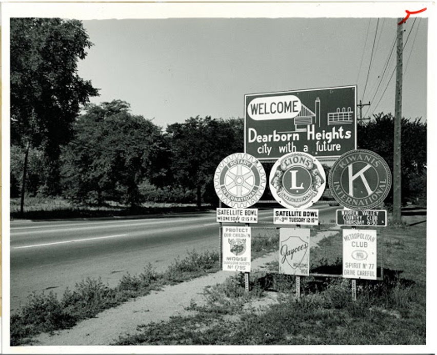 A photograph shows a road in the background with several street signs in the foreground. The most prominent being - Welcome to Dearborn Heights.