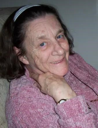 An elderly Bloetscher looks into the camera from the side. She is wearing a pink sweater and has a large smile on her face.