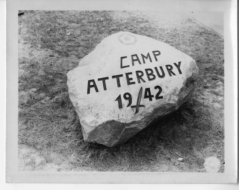 A black and white photograph of a large rock on grass that has been carved with the words “Camp Atterbury 1942.” In between the 19 and the 42 is an engraving of a sword.
