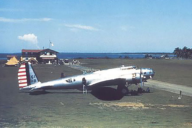 An airplane is in the foreground of the image, parked next to an airstrip. In the left background is a two-story building.