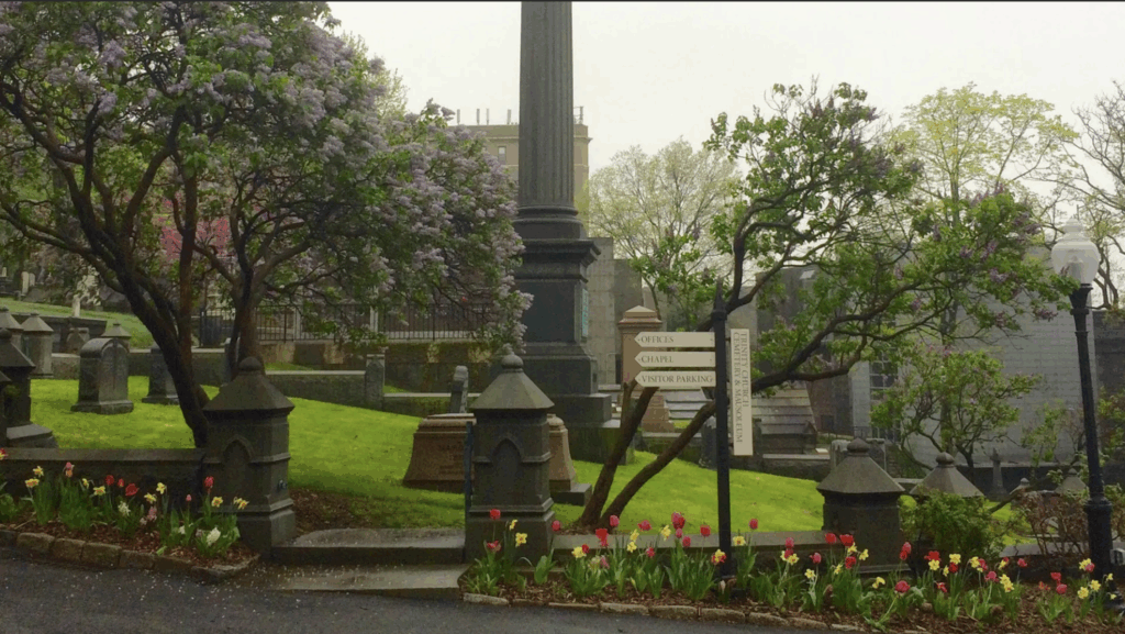 Closeup photograph of a cemetery with a monument, several graves, and a few trees.