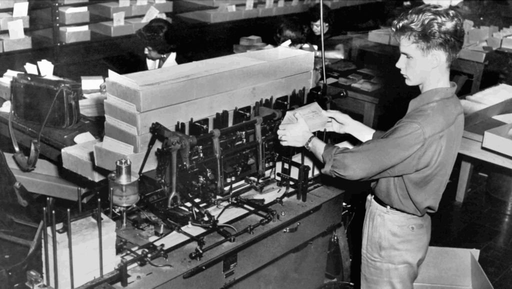 Photo of a machine with a man standing in front feeding envelopes.