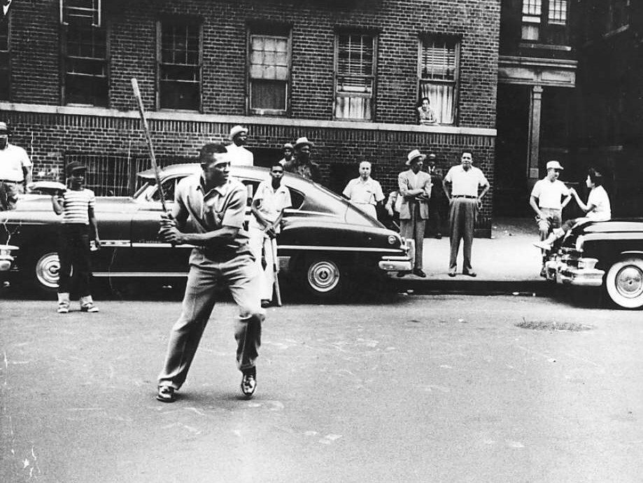 A photograph of Willie Mays playing stickball on the street with a car and a crowd of kids watching in the background.
