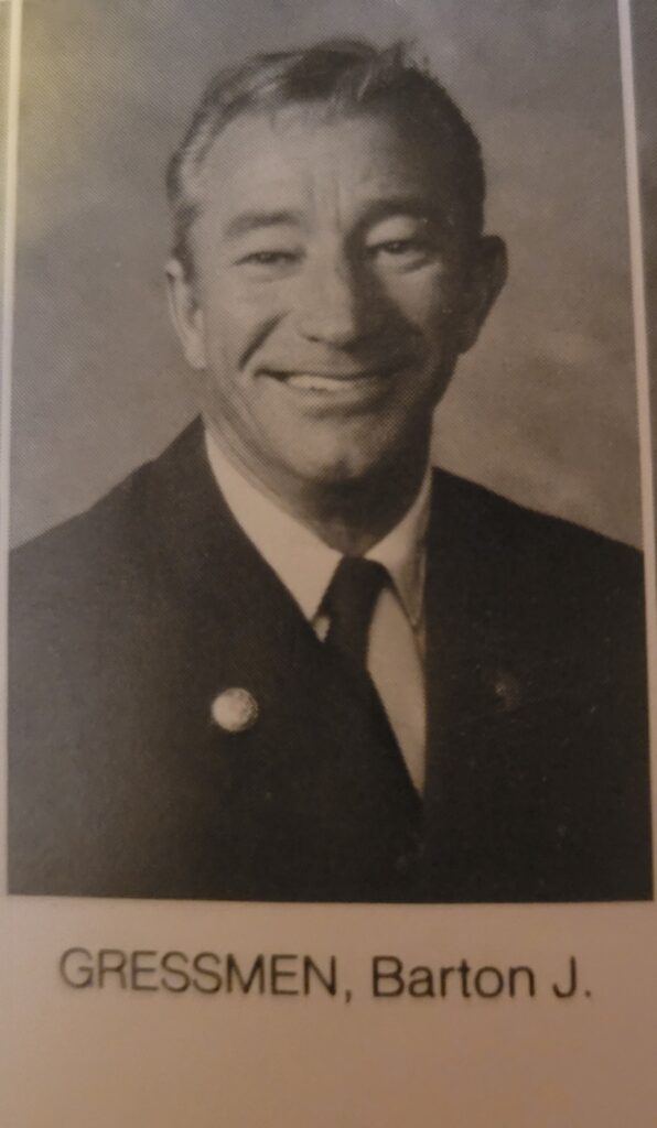 An older man smiles into the camera. He is wearing a suit and tie.