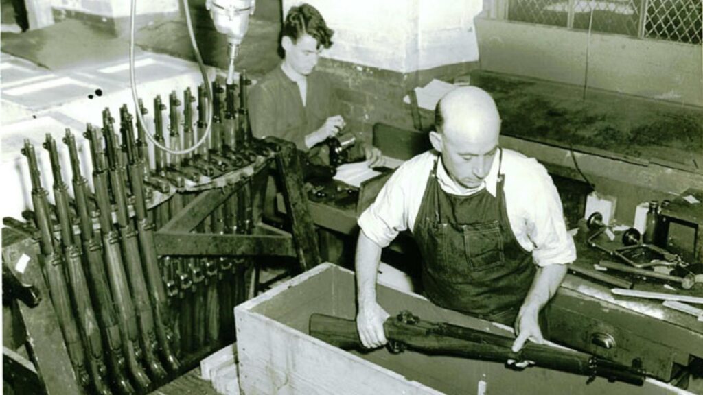 A black and white photograph of a factory with a man placing a gun in a box.