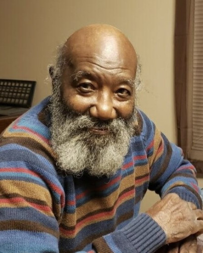 An older African American man looking into the camera. He is bald on the top with gray hair on the sides and a gray beard. He is sitting at a table in a house and wearing a striped sweater.