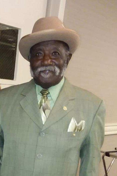 An elderly African American man is dressed in a green suitjacket with a light green shirt and metallic green tie and matching pocket square. He is wearing a tan fedora.