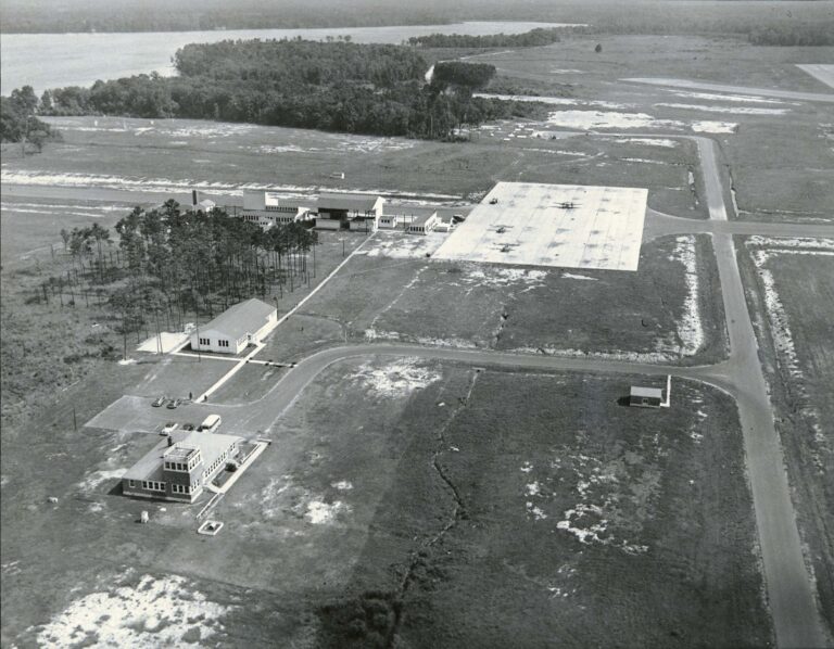 An aerial black and white photograph of an airstrip, landing pad with a few planes parked, and a few training buildings. The facility is situated in the middle of an open field with minimal tree coverage.