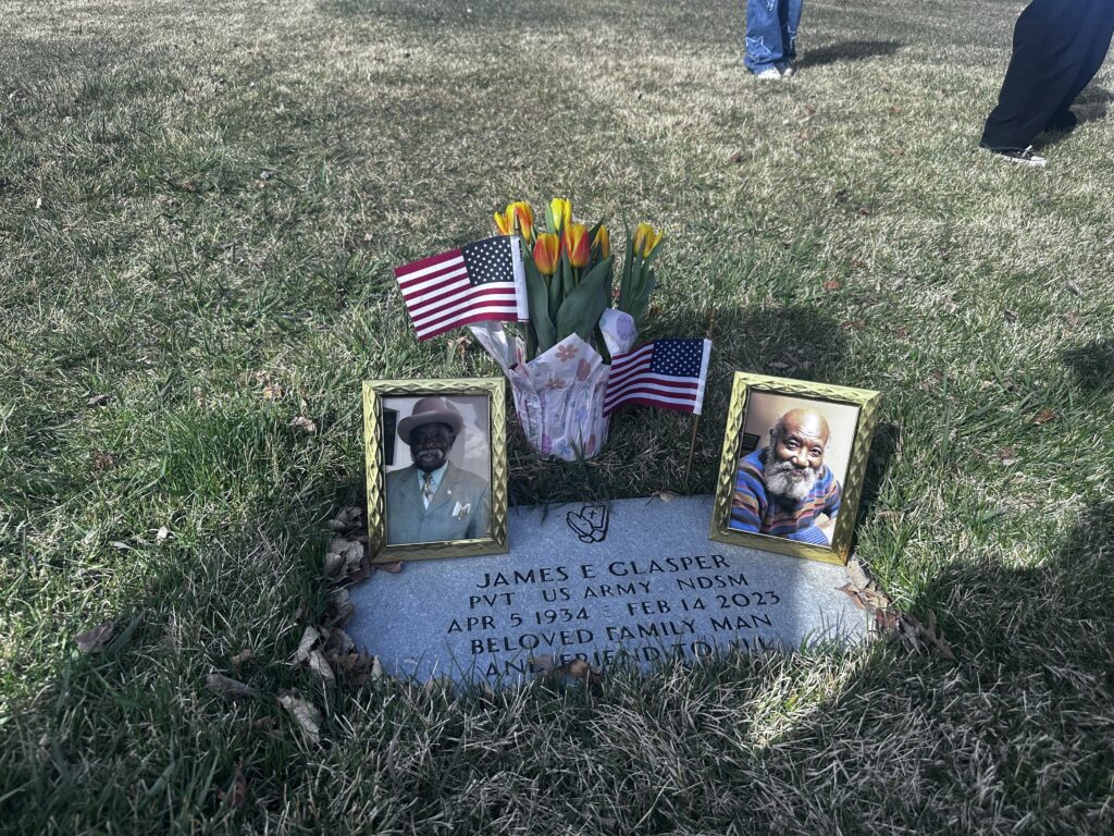 A flat marker is engraved with “James E Glasper PVT US Army NDSM APR 5 1934 FEB 14 2023 Beloved Family Man and Friend to All.” Around the grave are two framed photos of Glasper that were included in the above profile, two American flags, and a tulip plant.