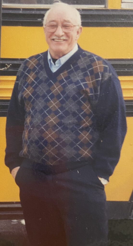 An older man in a sweater and dress slacks stands in front of a yellow school bus. He is wearing glasses and has a mustache and white hair. He is smiling toward the camera.