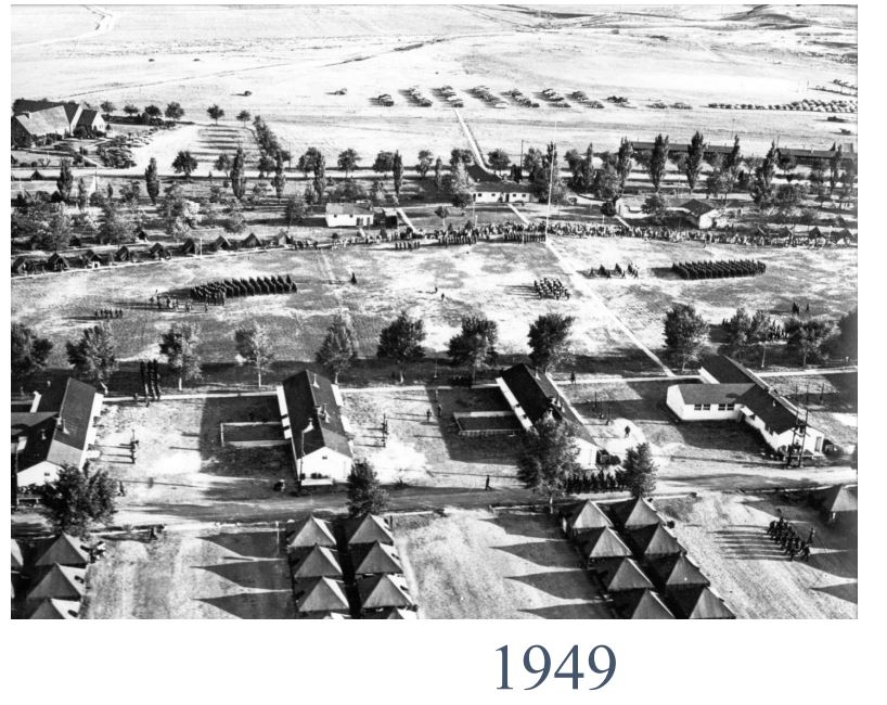 A black and white aerial photograph of multiple barracks on lined streets. There are several companies of soldiers standing in formation throughout the image.