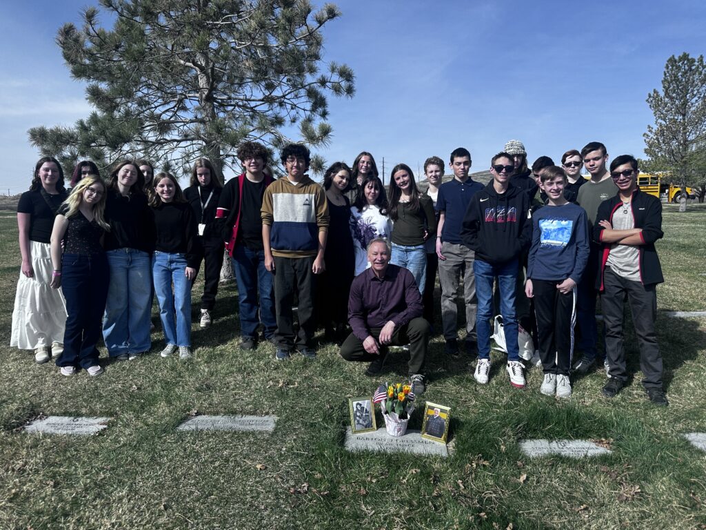 Several students stand around Gressmen’s grave marker. His son is an older man and is kneeling in front of the students and behind his father’s marker.