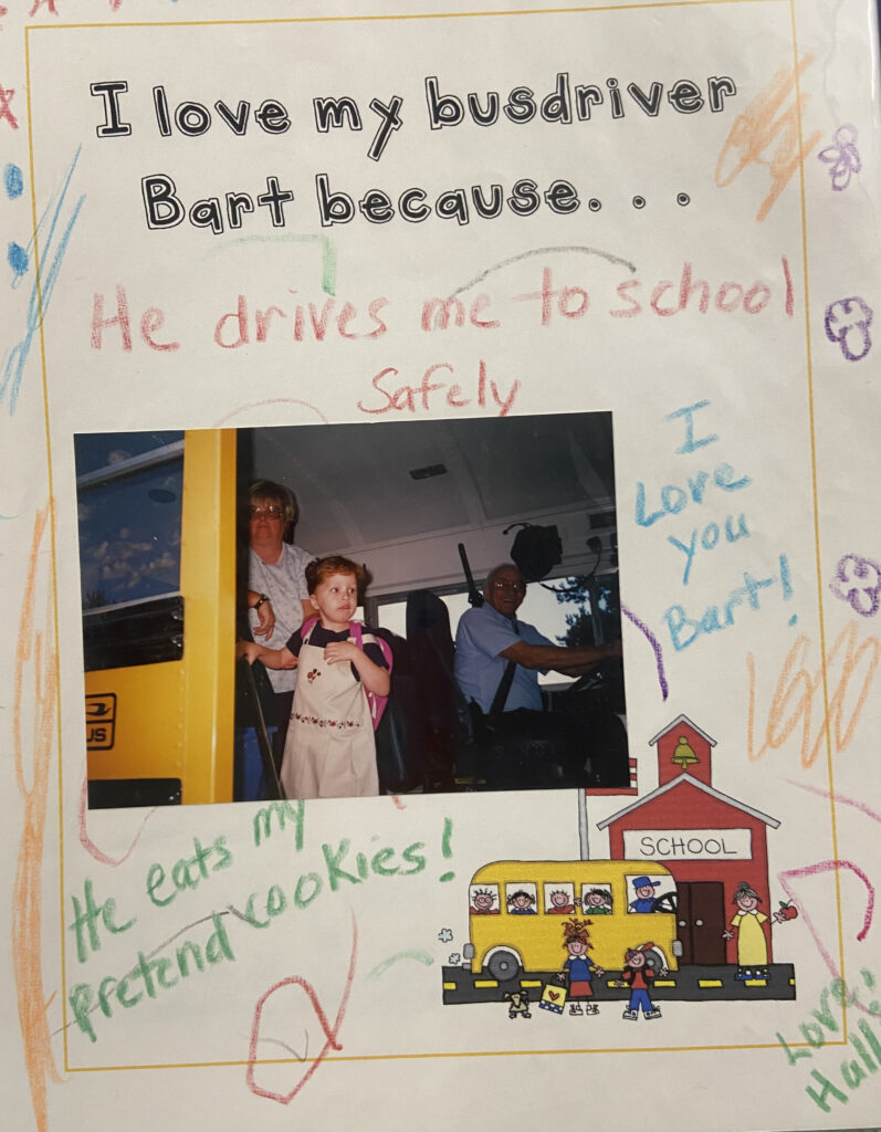 A student worksheet with the words ‘I love my busdriver Bart because…” typed at the top. Around it in crayon are the words, “He drives me to school safely, I love you Bart!, and He eats my pretend cookies.” In the center of the worksheet is a photo of a young student getting off the bus with a pink backpack. There is an older woman bus aide standing behind the child and Bart is sitting in the driver’s seat of the bus, smiling toward the camera.