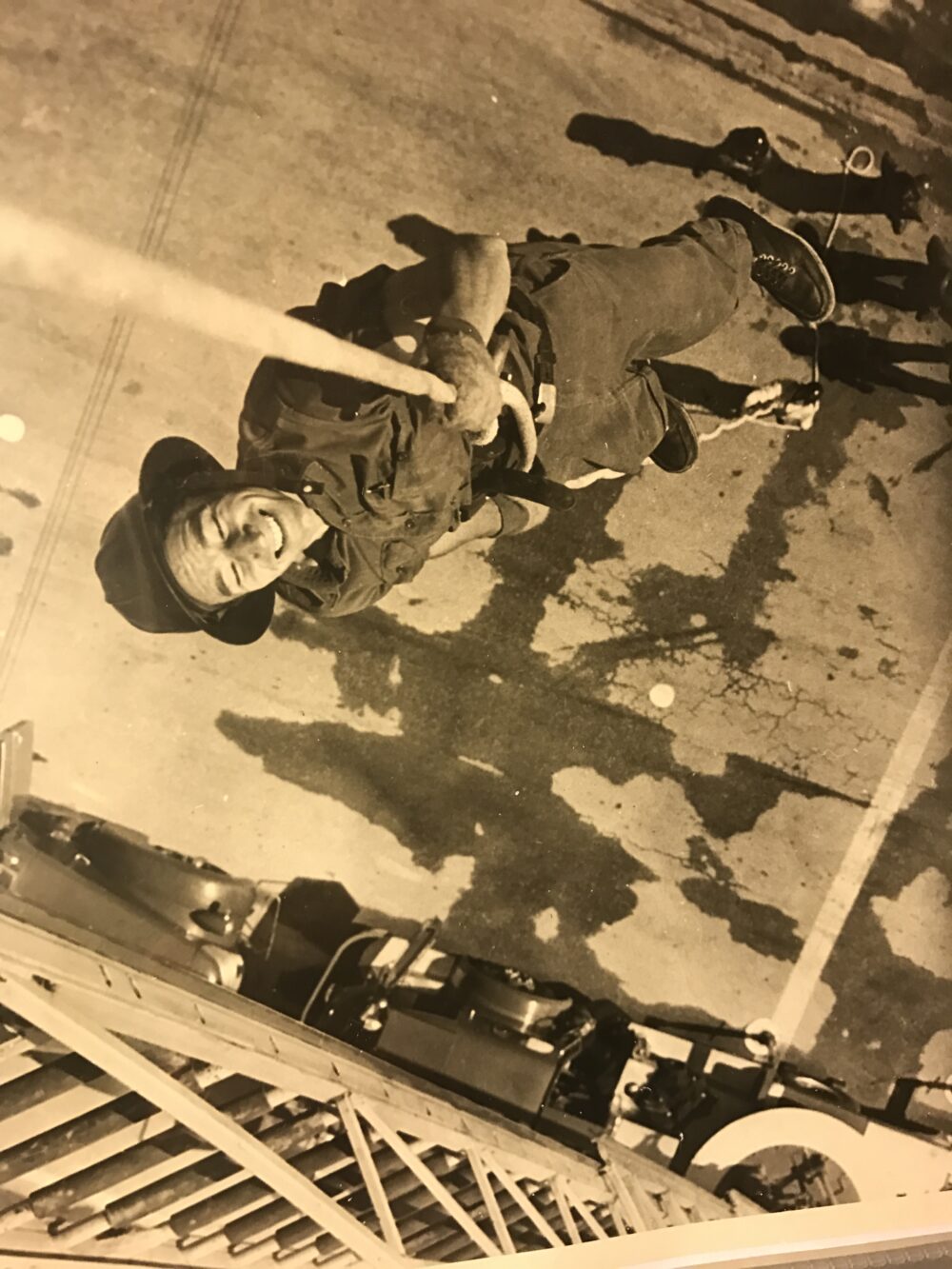 A sepia photo of a man in work pants and rolled up sleeves of a workshirt, along with a wide-brimmed hat, spraying a firehose into the air and smiling with his eyes closed.