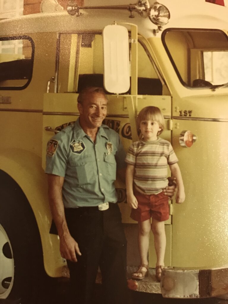 An older man is wearing a uniform with a short-sleeved blue shirt and black pants. A young boy stands on a yellow firetruck next to him. The boy is wearing red shorts and a striped shirt. Both are smiling in the camera.