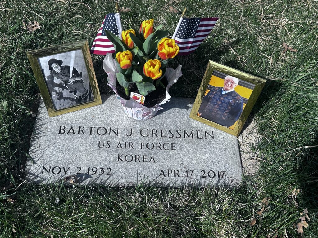 A flat, rectangular, marble marker is engraved with, “Barton J. Gressmen US Air Force Korea Nov 2 1932 Apr 17 2017.” On the left-side of the marker is a picture of Gressmen as a firefighter. On the right, is a photo of him in front of his bus. Both photos are in gold frames. In the middle is a tulip plant with two American flags.