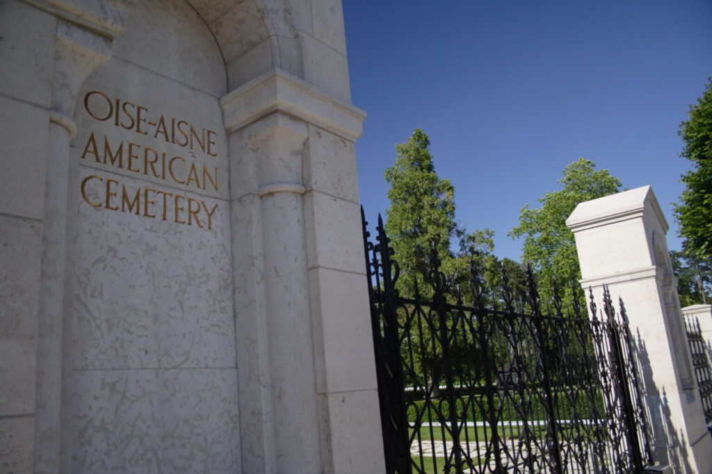Two peaked marble arches are connected via a iron fence. On the left-most arch reads ‘Oise-Aisne American Cemetery’