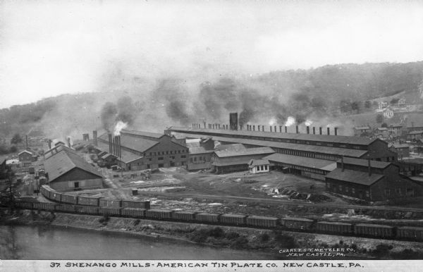 A sprawling mill sits on the bank of a river. Along the river runs a trailway. In the distance is a small town of houses, followed by woods.