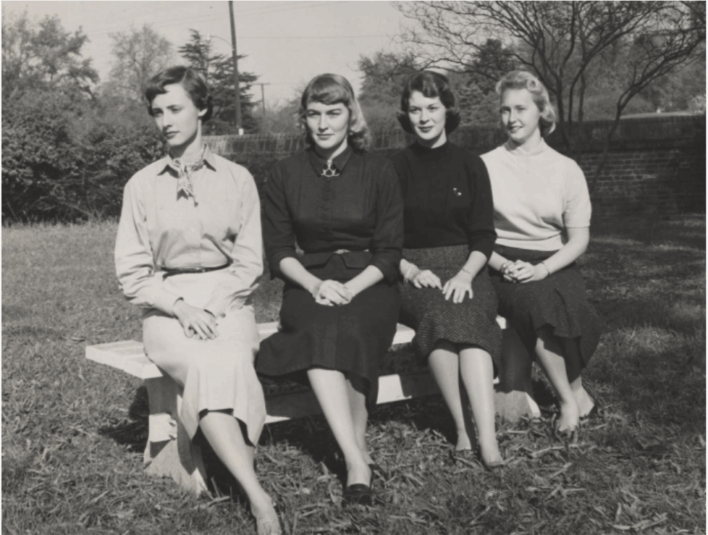 Four young women are sitting on a bench in a fenced-in yard. They are all dressed in skirts and blouses.