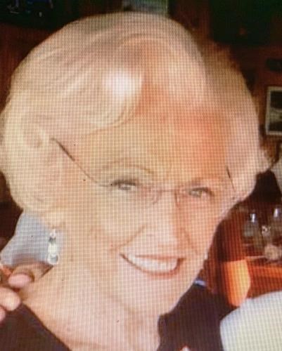 An older woman with short, blonde hair and glasses is smiling into the camera.