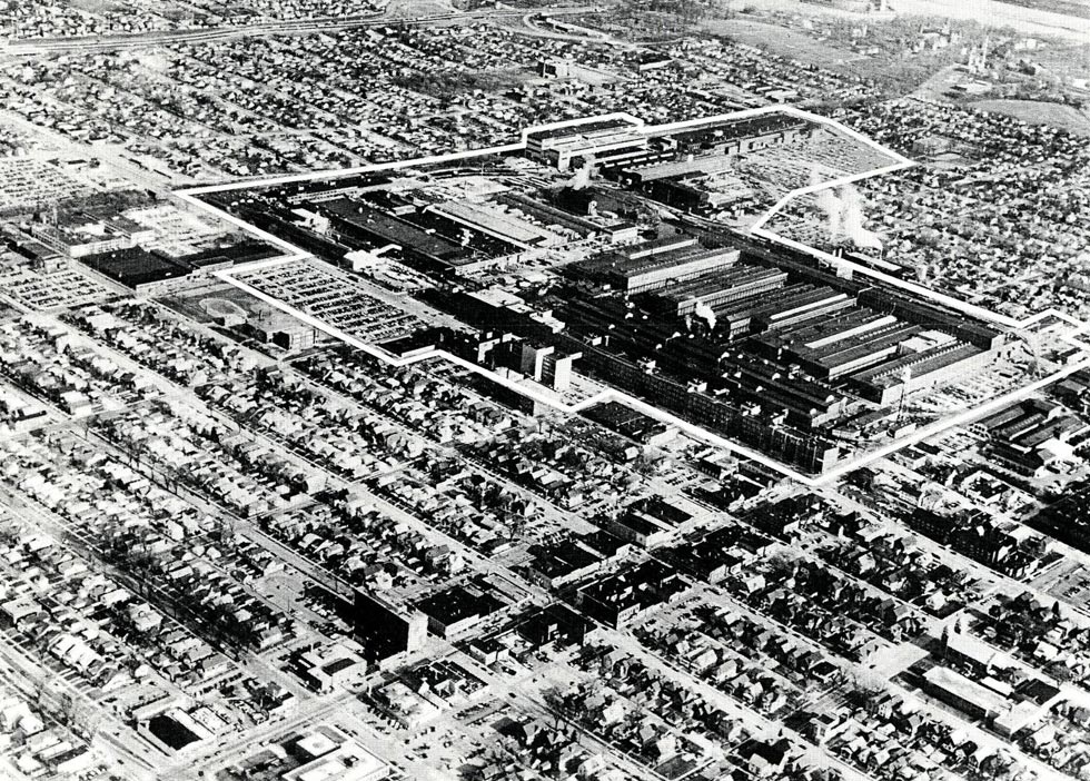 A black and white aerial image of a massive industrial complex in the middle of the city with houses surrounding it.