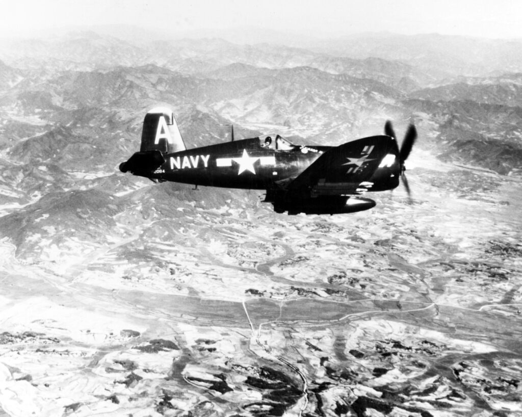 A black and white aerial image of an open cockpit plane in flight over mountainous terrain.