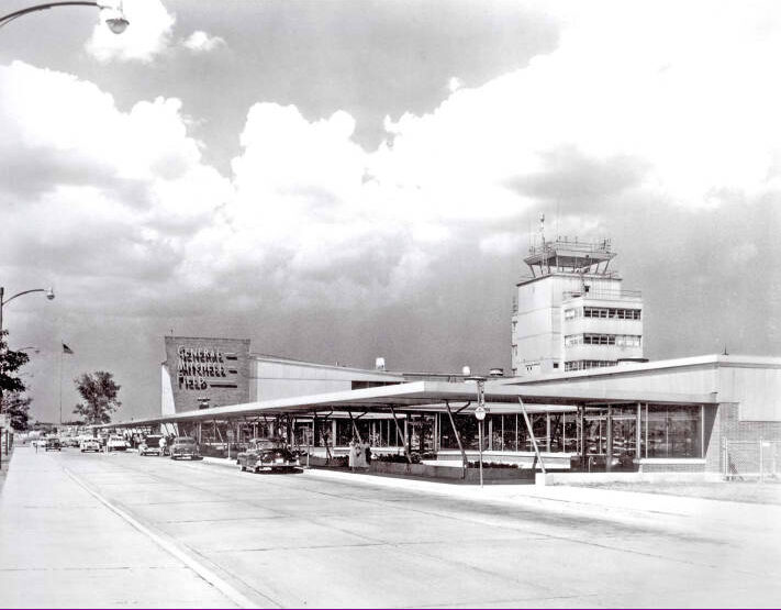 A black and white image of a outdoor canopy attached to the airport terminal. There are several 1950s cars parked at the surb. In the building on the distance it says, “General Mitchell Field.”