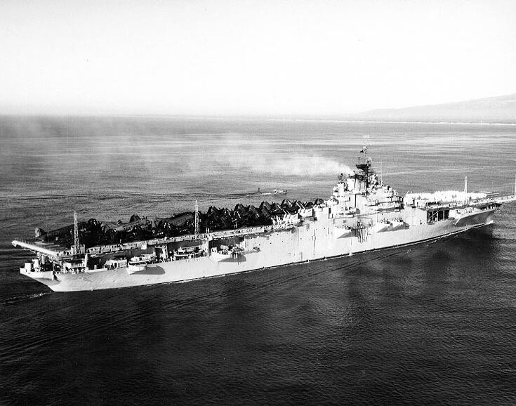 A black and white aerial image of a large ship full of airplanes in the middle of the ocean. The shore can be seen in the far distance.