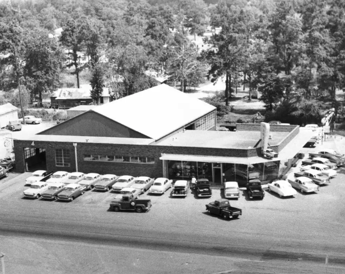 A black and white aerial image of a car dealership building. On the right is a glassed showroom, and on the left is a large attached garage. Outside, there are several new vehicles parked.