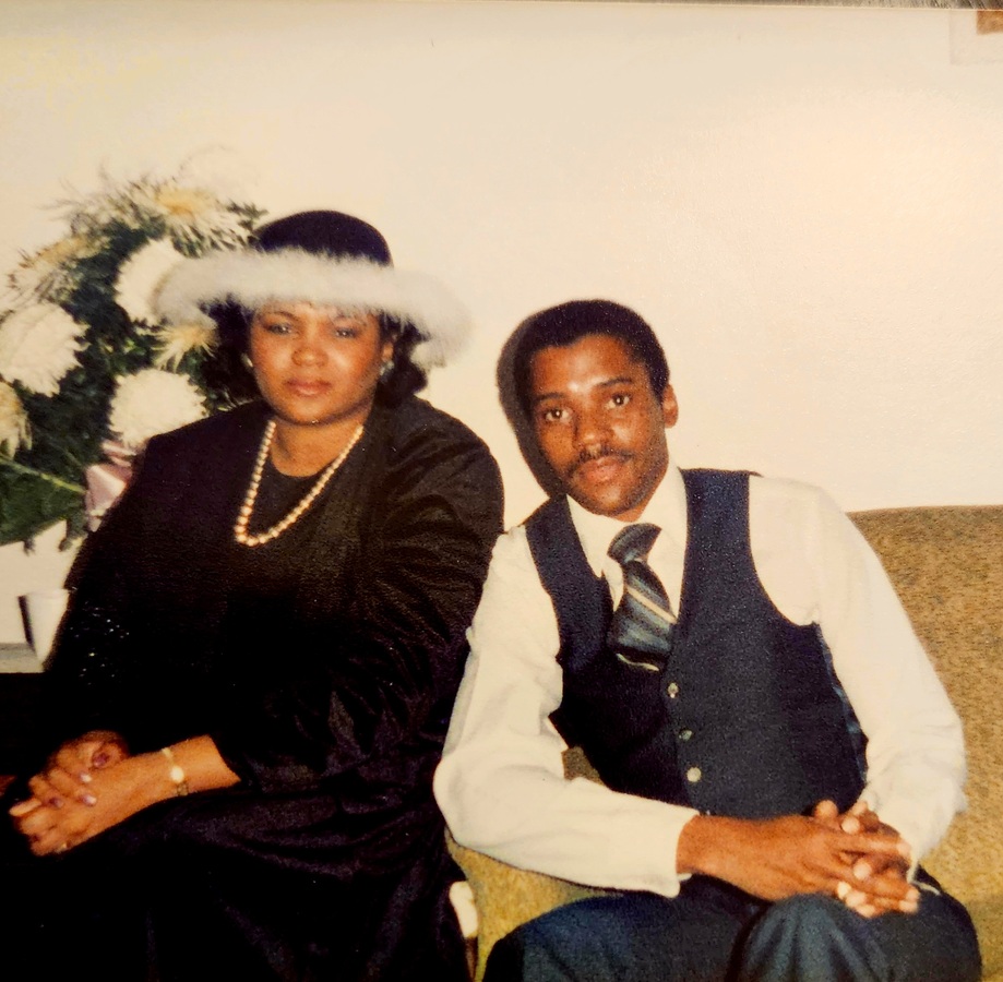 A color image of a young Black couple. The woman on the left is wearing a black dress with a black hat trimmed in white fur. She has a pearl necklace on. The man is in a suit vest and pants with a matching blue striped tie and white dress shirt. They are sitting next to each other.