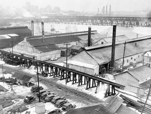 A black and white aerial image of an abandoned train trestle and several warehouses. There are other bridges, smokestacks, and a city line in the distance.