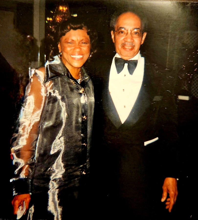 An older Black couple is standing next to each other. They are in formal attire and smiling at the camera.
