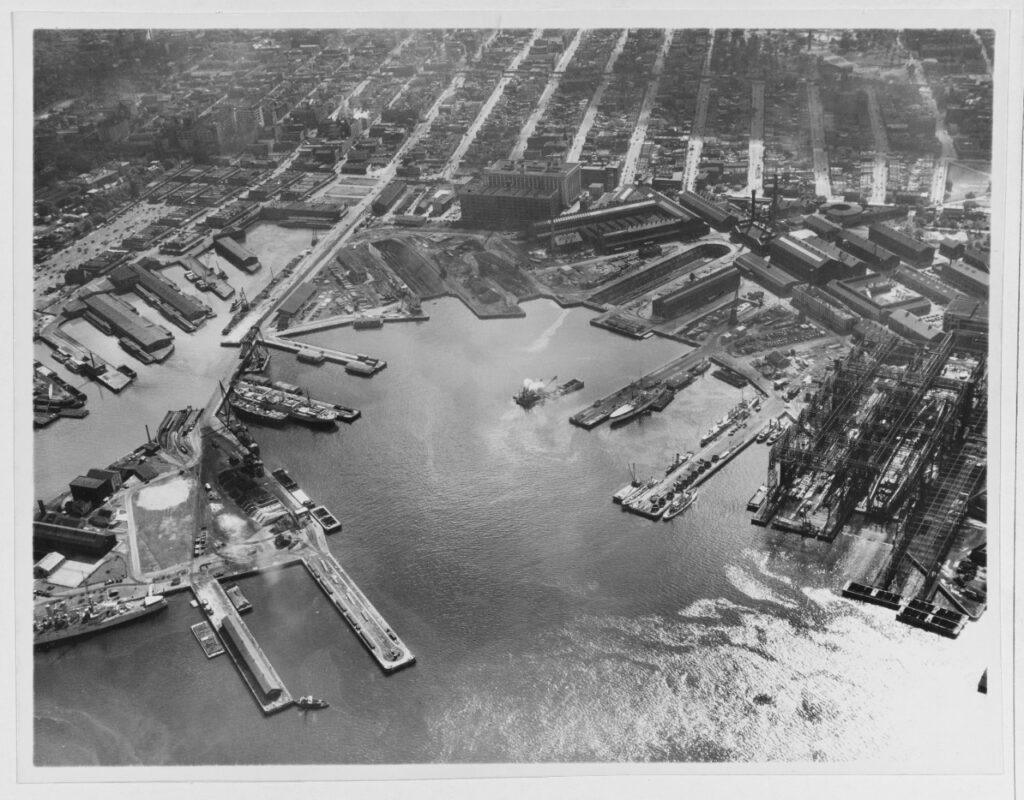 A black and white aerial image of several boat docks. To the right there are a few ships being built under scaffolding.