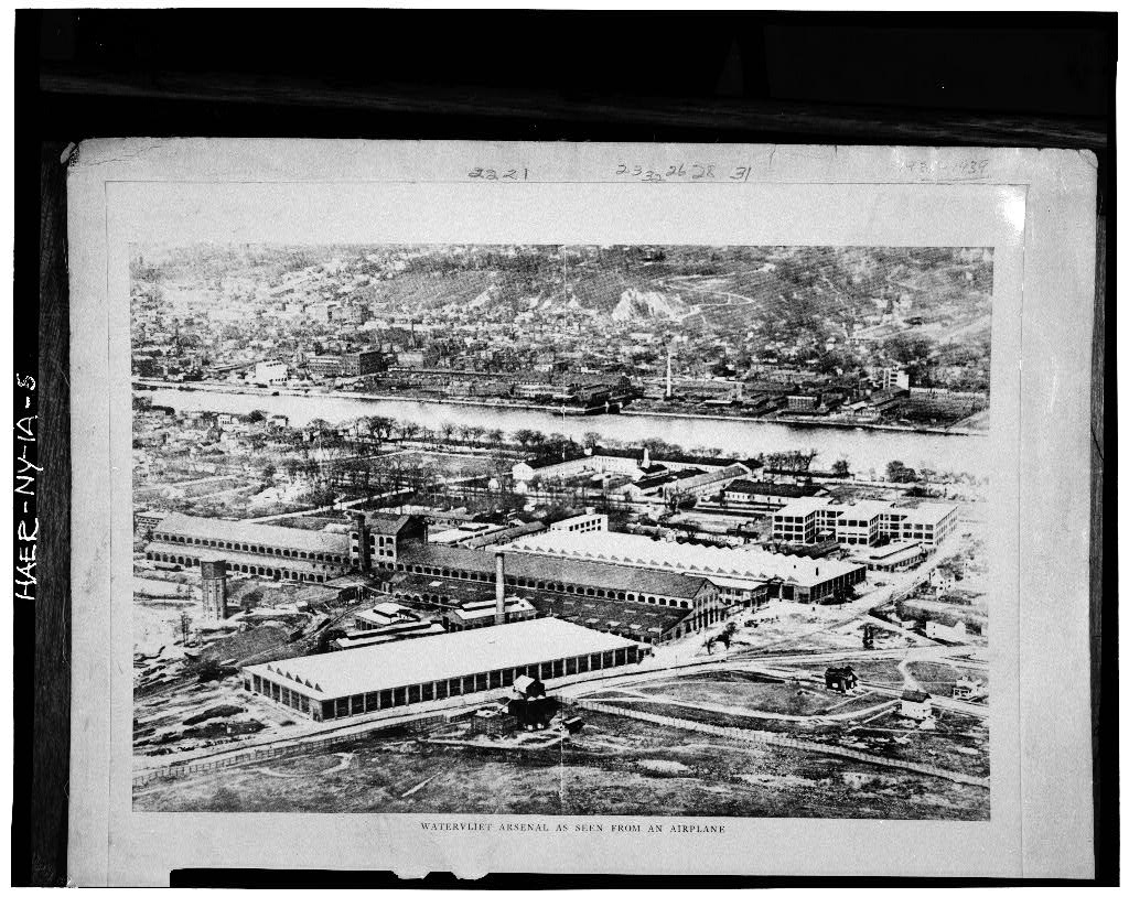 A black and white aerial image of several large factory-type buildings on the banks of a river.