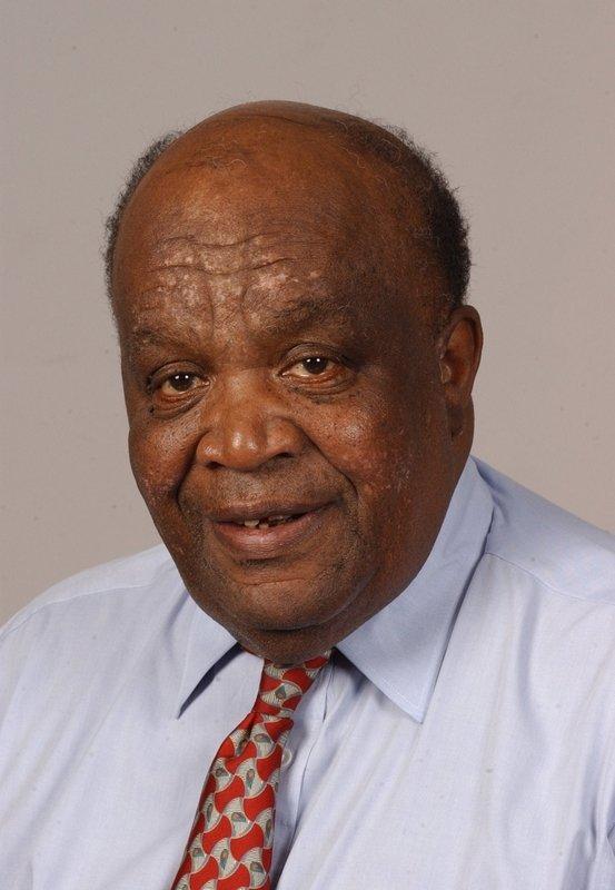 An older African American man who is bald on top looks directly into the camera while smiling. He is wearing a light purple button-down shirt and a red and gray patterned tie.