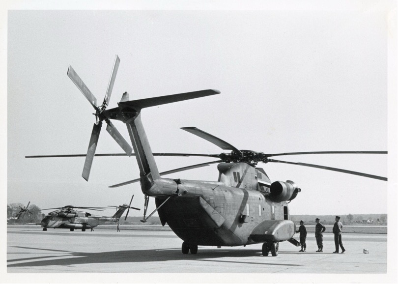 A black and white photograph of a large helicopter on the ground. Three men are standing outside the door looking in. There are a few other helicopters in the background.