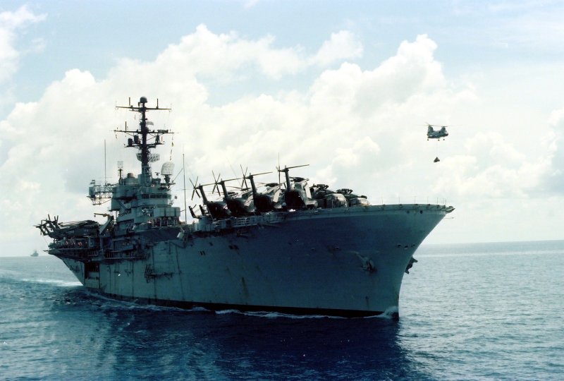 A color image of a ship in the middle of the ocean. On the deck are several helicopters. One helicopter is in the sky near the ship.
