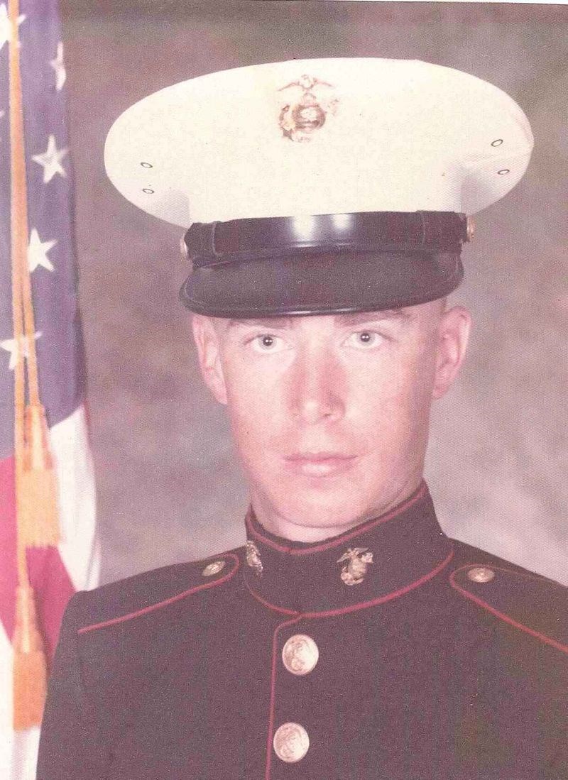 A headshot of a young, White man in full Marine dress uniform. There is an American flag behind his right shoulder.