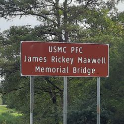 A brown highway sign reading “USMC PFC James Rickey Maxwell Memorial Bridge”