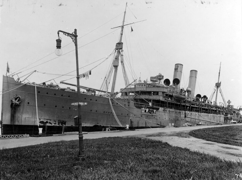 A black and white image of a large ship tied up along a sidewalk.