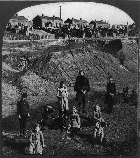 Stereograph black and white image showing a group of children in foreground; spoil tips and wooden framed houses in background.
