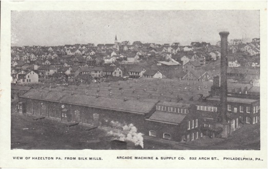 A black and white image of an aerial view of a town. In the foreground are many factory buildings in a row with a large smokestack. In the background are dozens of homes.