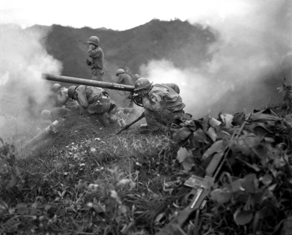 A black and white image of soldiers covering their ears while 75mm guns are going off. 