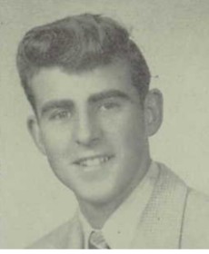 A black and white headshot image of a young man looking directly into the camera. He is wearing a suit and tie and is smiling.