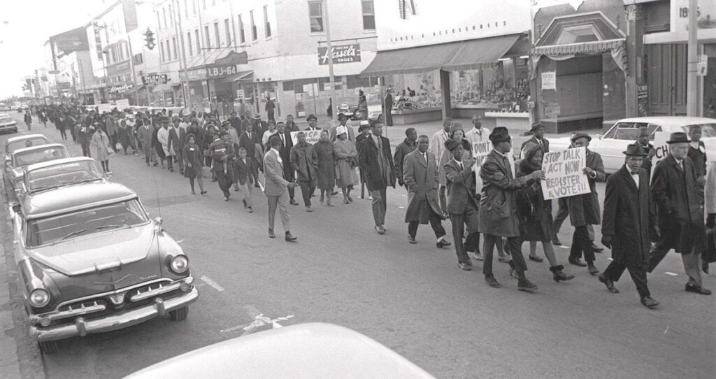 Dozens of protestors walk down the middle of a street holding signs.