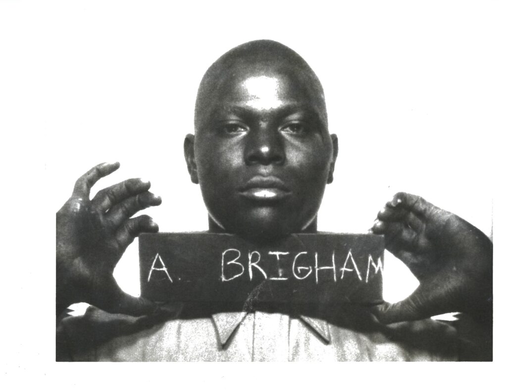 A black and white photo of a young African American man holding a sign under his chin that says 'A. Brigham.'