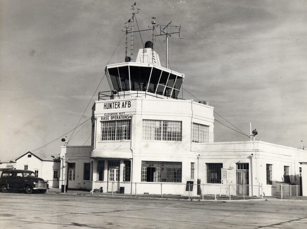 A black and white photo of a base operation building. The middle part of the building is two stories with an observation tower on top and many antennas.