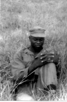 A Black and white image of an African-American man sitting in a field in uniform. He has one leg bent and his hands are wrapped around it.