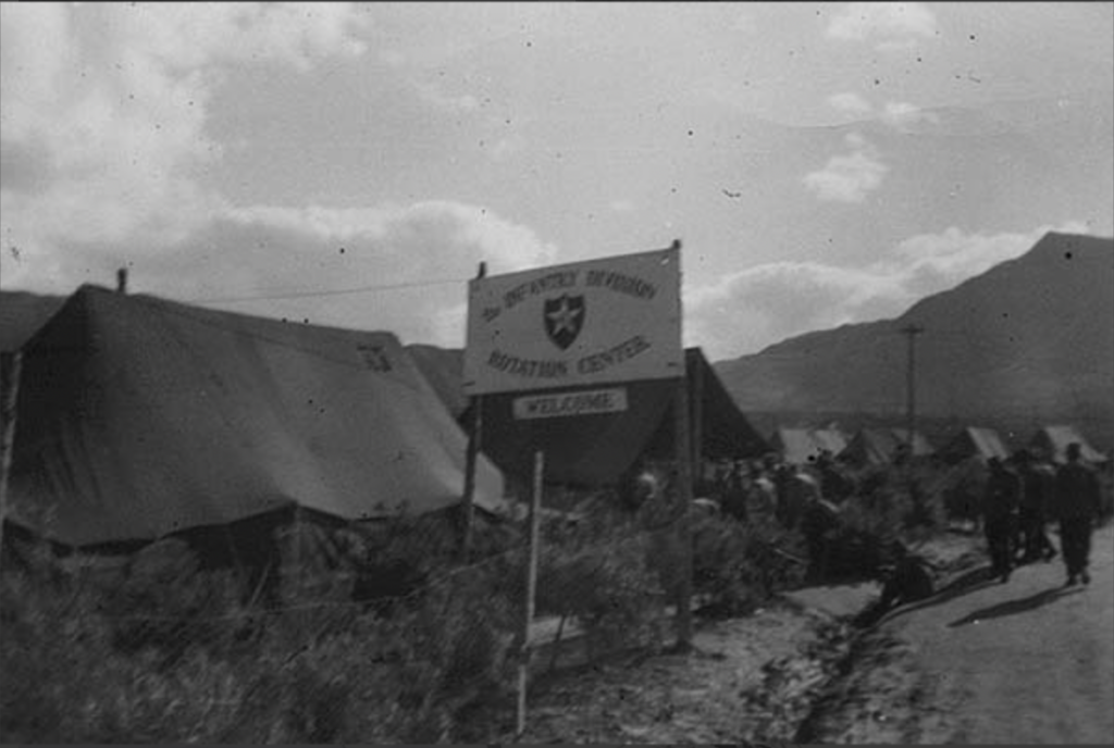 A black and white photo shows several tents set up in the background. In the front is a sign that says ‘2nd Infantry Division Rotation Center Welcome’. In the back are mountains.