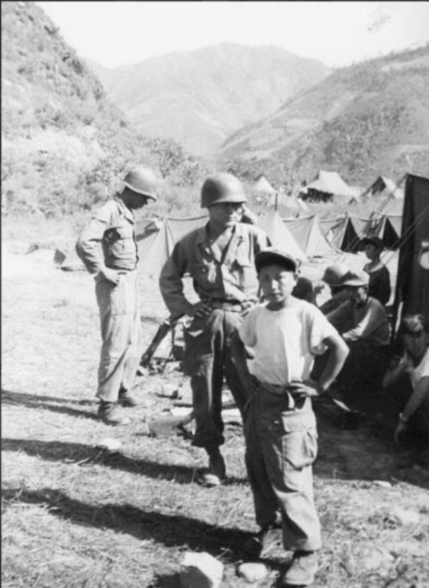 A black and white photo of a young Korean boy standing and looking at the camera with his hands on his hips. Behind him are several U.S. soldiers both sitting and standing. There are several tents around and mountains in the background.